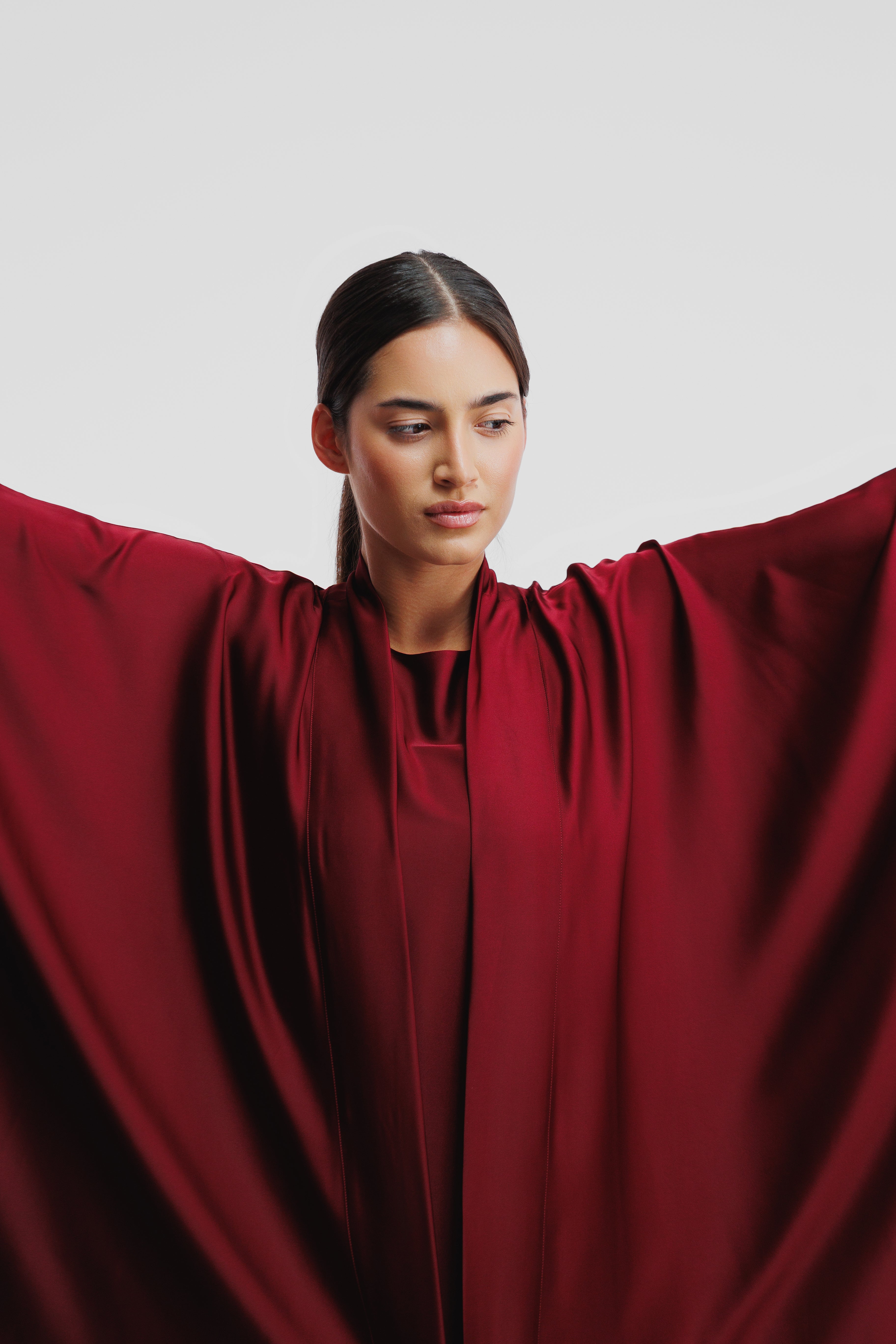 A woman wearing a flowing deep burgundy cape-style top with wide draped sleeves, posed against a plain light gray studio background.