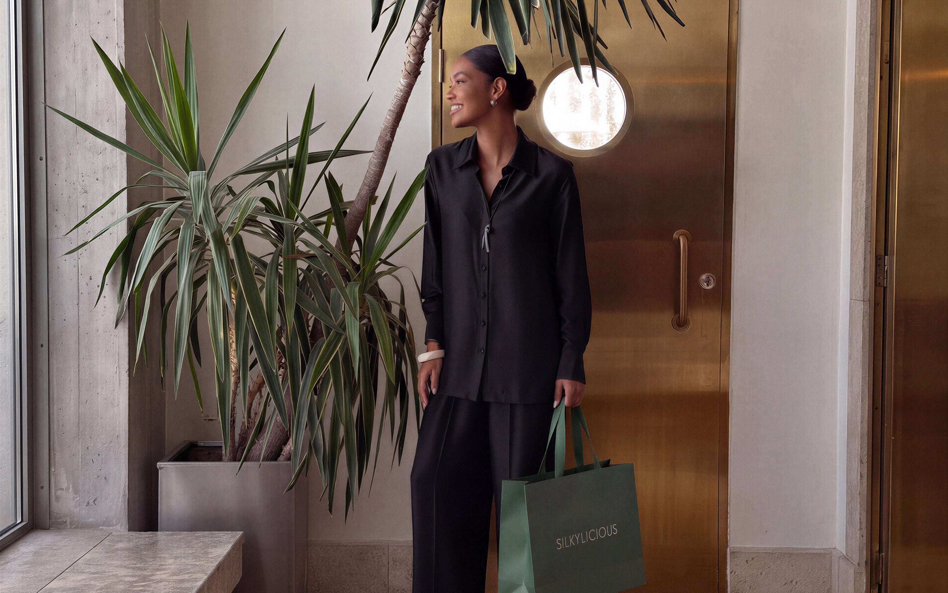 A woman wearing a black button-up shirt and matching black trousers stands indoors next to a large potted plant, holding a green shopping bag with the brand name 'SILKYLICIOUS' printed on it.